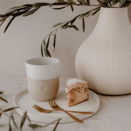 Beige keramische koffiemok met hartdetail op bord met taartpunt naast minimalistische vaas